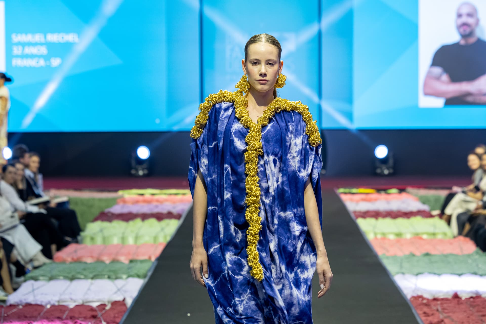 Vestido azul tie-dye com aplicações douradas em desfile de moda